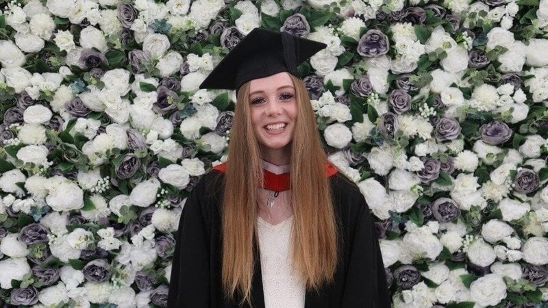 Iesha Iesha graduating in front of a wall of flowers